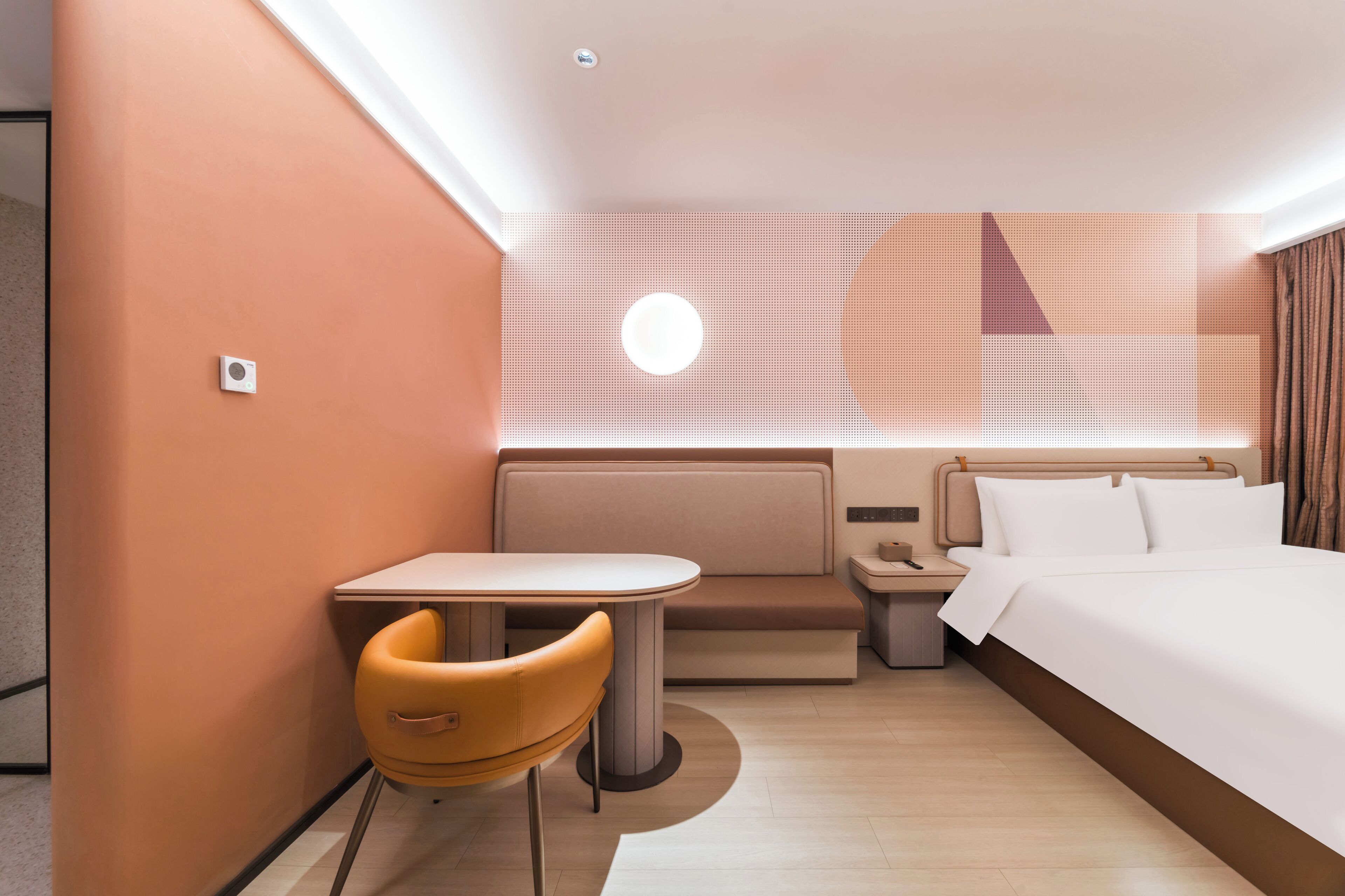 A modern hotel room with a bed, a small table with a chair, a nightstand, and a wall-mounted light.