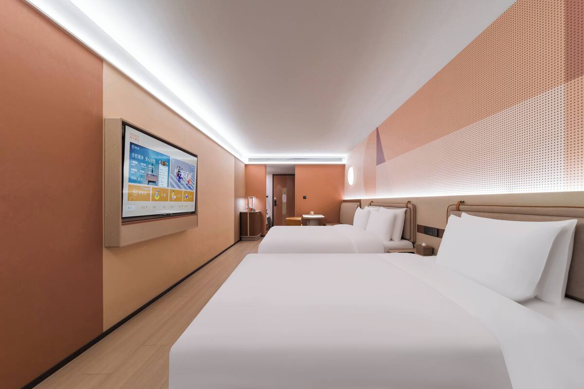 Una habitación de hotel con dos camas, un televisor montado en la pared y una mesita con una lámpara.