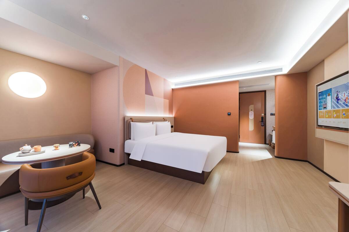 Una habitación de hotel moderna con una cama, una mesa redonda con una silla, una mesita con un calentador de agua y tazas, y un televisor montado en la pared.