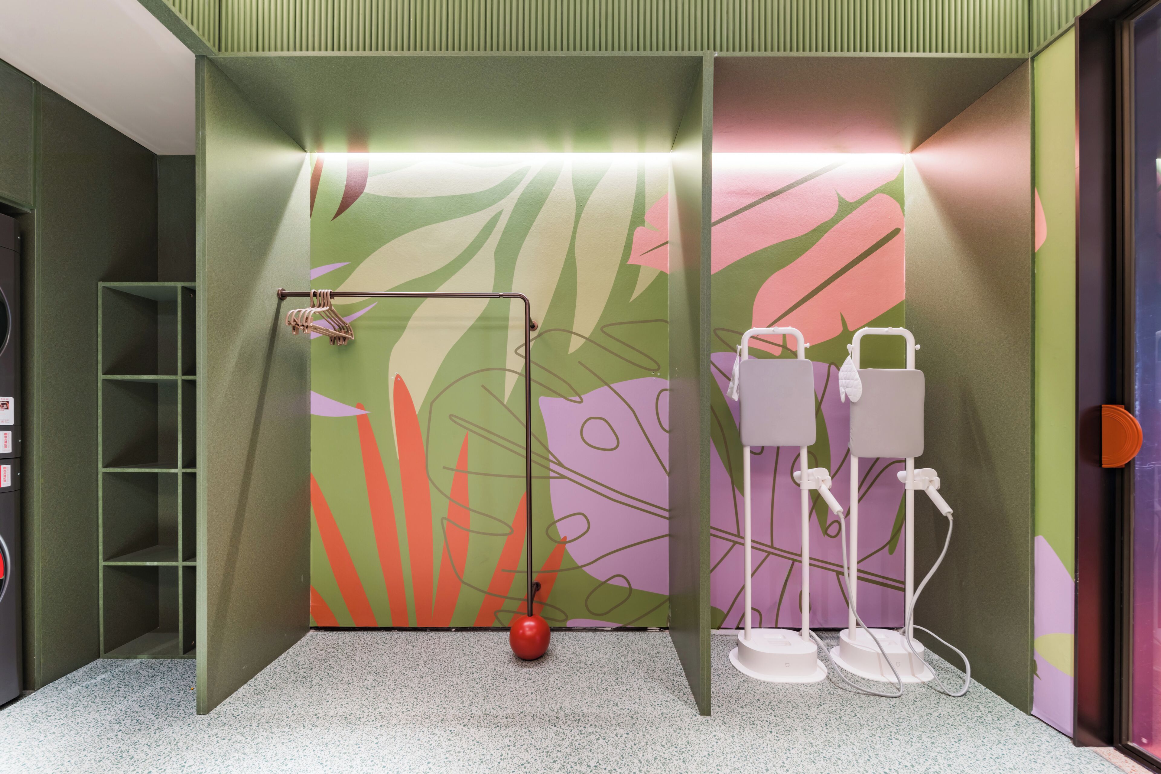 A bathroom with a green and pink leaf-patterned wall, a red hanging towel rack, and two white towel dispensers.
