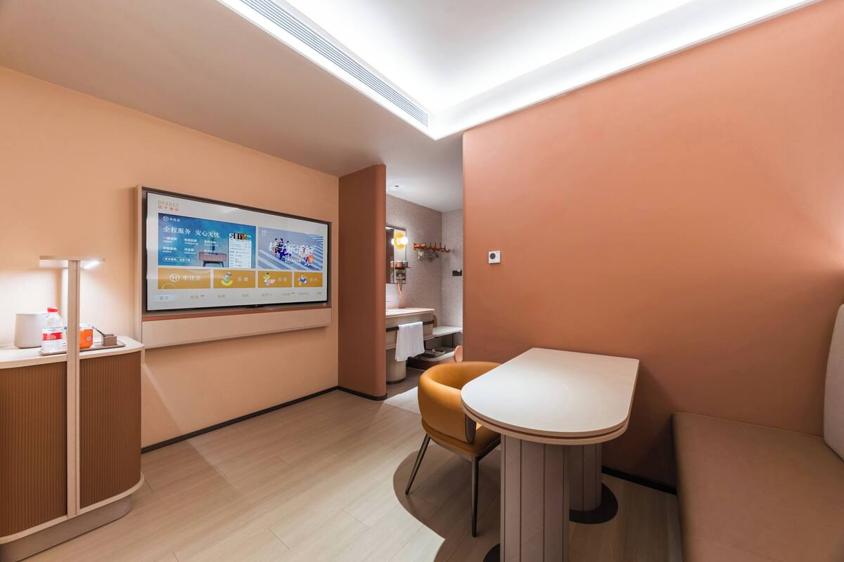 Una habitación de hotel moderna con una pantalla digital grande, una mesa redonda, un sofá y una zona de baño pequeña.