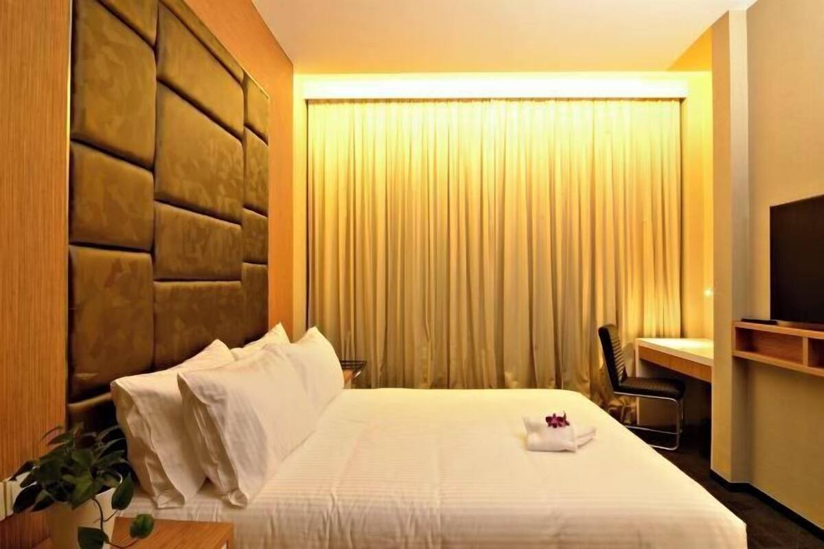 Une chambre d’hôtel avec un grand lit, une tête de lit, un bureau avec une chaise, une télévision et une fenêtre avec des rideaux.