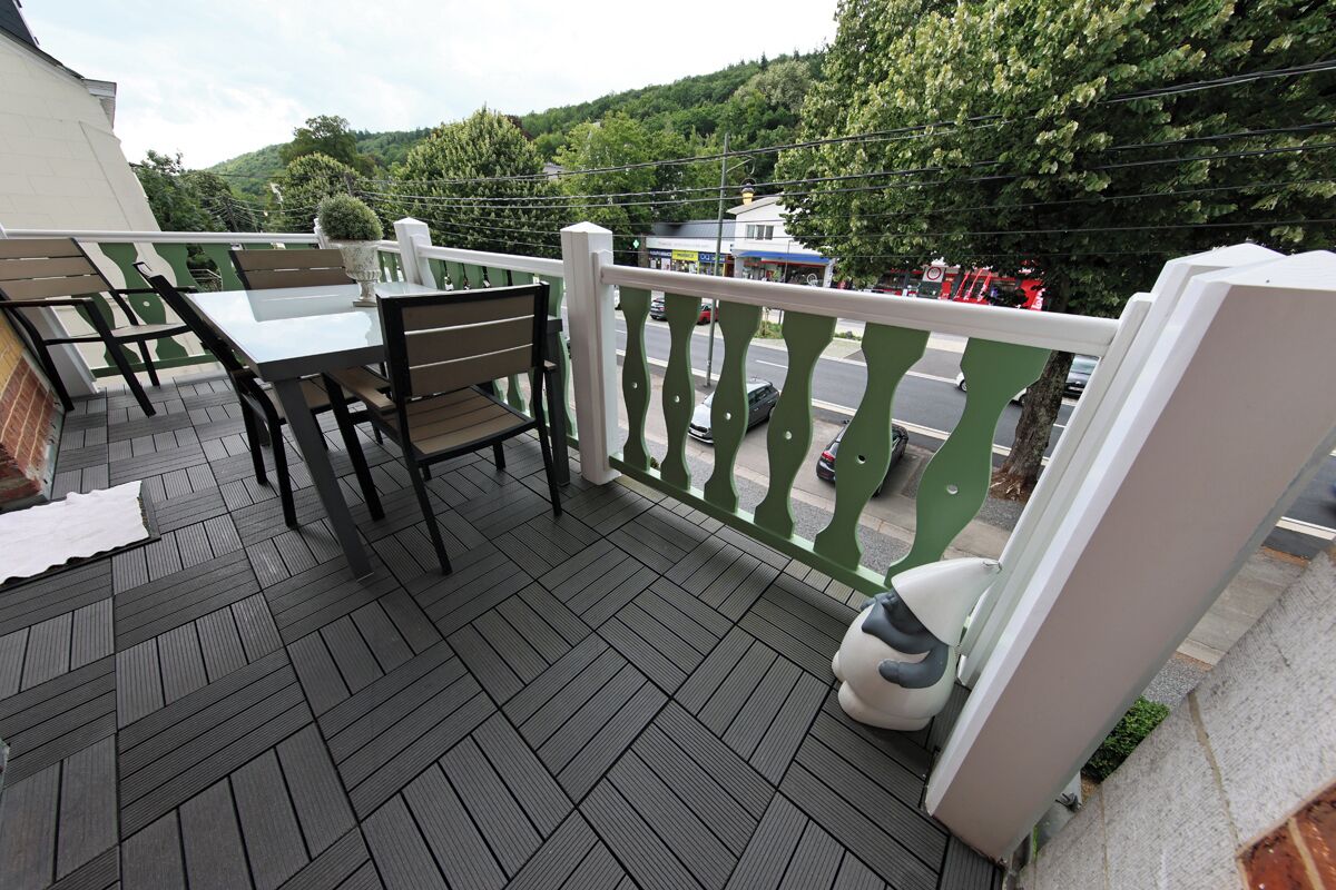 Een balkon met een tafel en stoelen, een witte leuning en een uitzicht op een weg en begroeiing op de achtergrond.