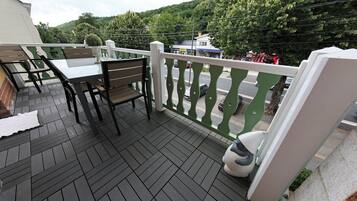 Een balkon met een tafel en stoelen, een witte leuning en een uitzicht op een weg en begroeiing op de achtergrond.