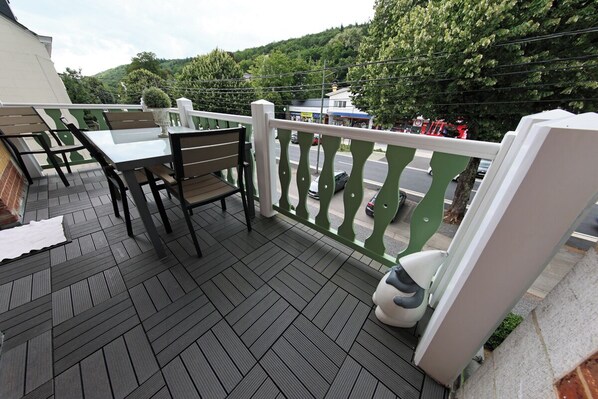 A balcony with a table and chairs, a white railing, and a view of a road and greenery in the background.