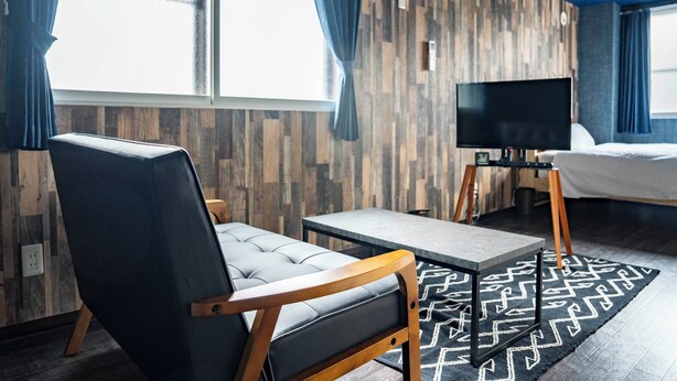Una habitación de hotel moderna con una pared decorada en madera, un sofá de cuero negro, una mesita, un televisor sobre un mueble bajo y una cama con alfombra estampada.