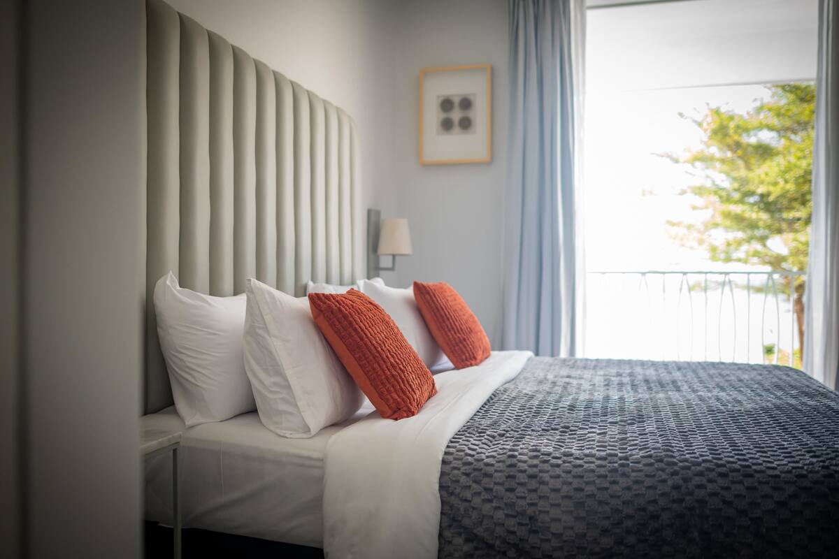 A hotel room with a bed, white and orange pillows, a grey patterned bedspread, a wall-mounted lamp, and a window with a view of trees.