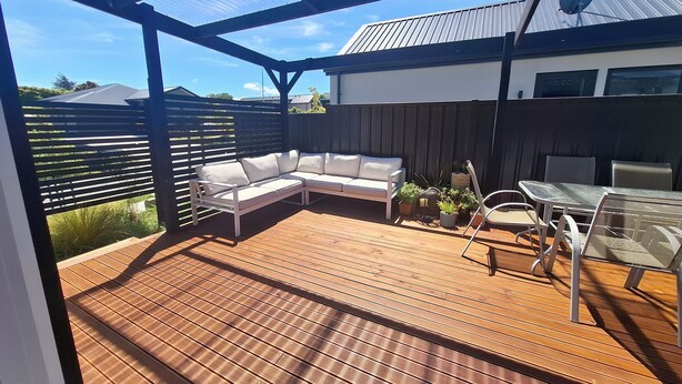 Eine überdachte Terrasse mit Holzdeck, einem weißen Ecksofa und einem Esstisch mit Stühlen.