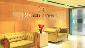 Een receptiegedeelte met twee groene banken, een houten bijzettafeltje met een plant in pot, en een muur met de naam 'MAALI & MANWIP'S' in metalen letters.
