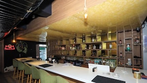 Een bar met een lange toonbank, groene barkrukken, planken vol flessen en een 'Red Bull'-bord.