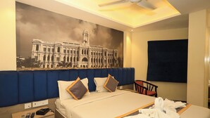 Een hotelkamer met een groot bed, een blauw hoofdbord, een plafondventilator, een aan de muur bevestigde televisie en een muurschildering van een historisch gebouw.