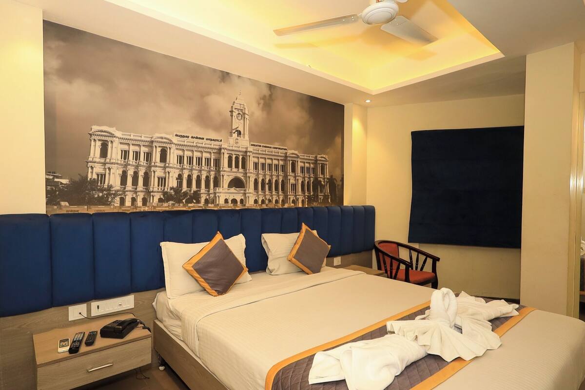 Een hotelkamer met een groot bed, een blauw hoofdbord, een plafondventilator, een aan de muur bevestigde televisie en een muurschildering van een historisch gebouw.
