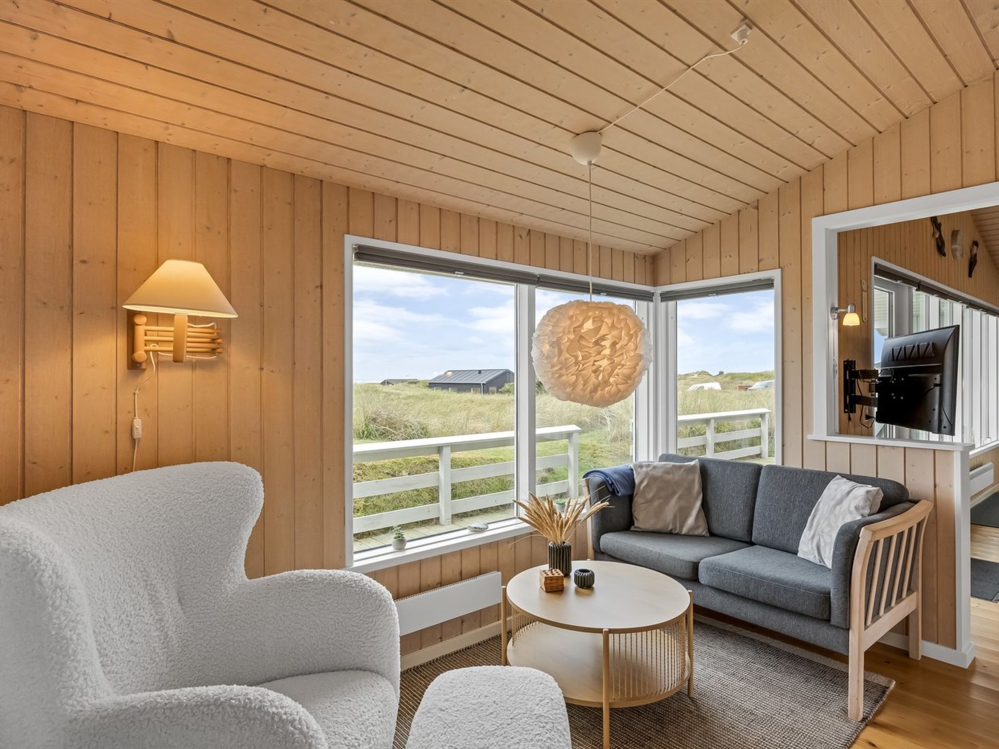 A cozy room with a wooden ceiling, a sofa, a chair, a round table, and a large window offering a view of the outdoors.
