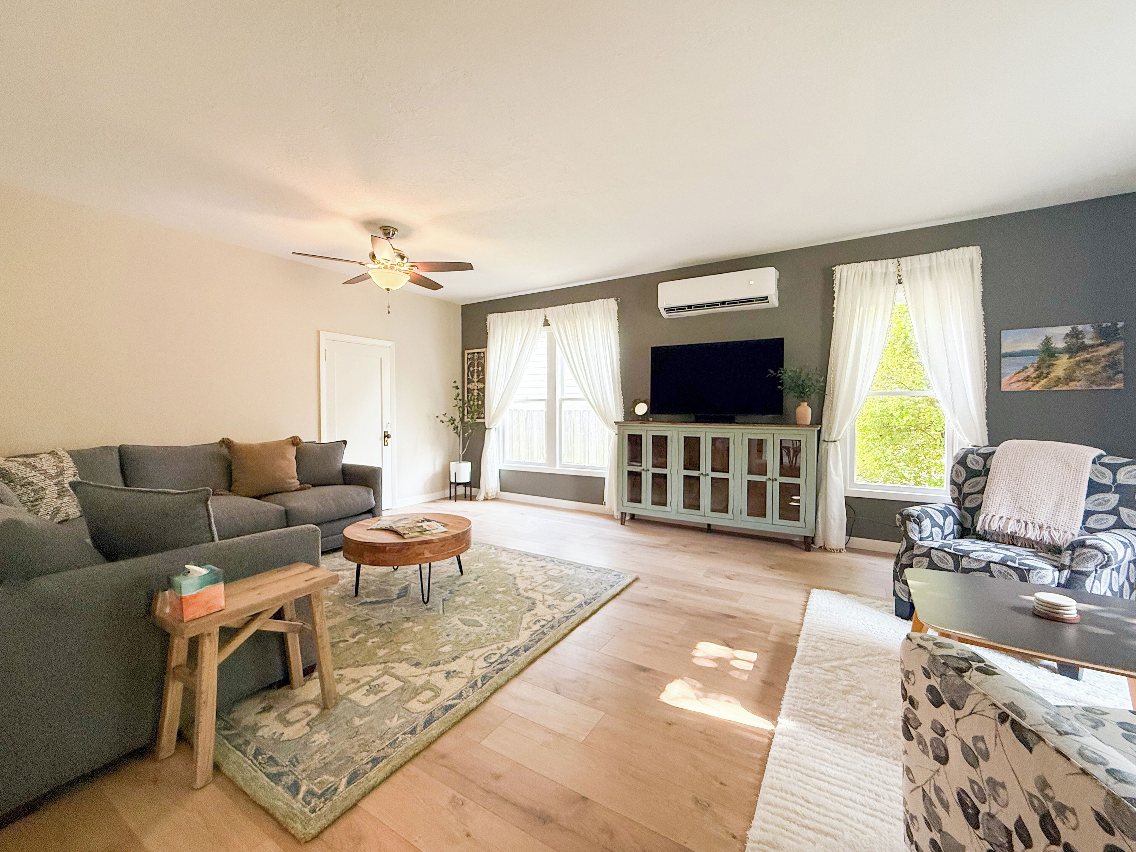 A living room with a grey sofa, a wooden coffee table, a patterned rug, a ceiling fan, a TV on a cabinet, and a window with curtains.