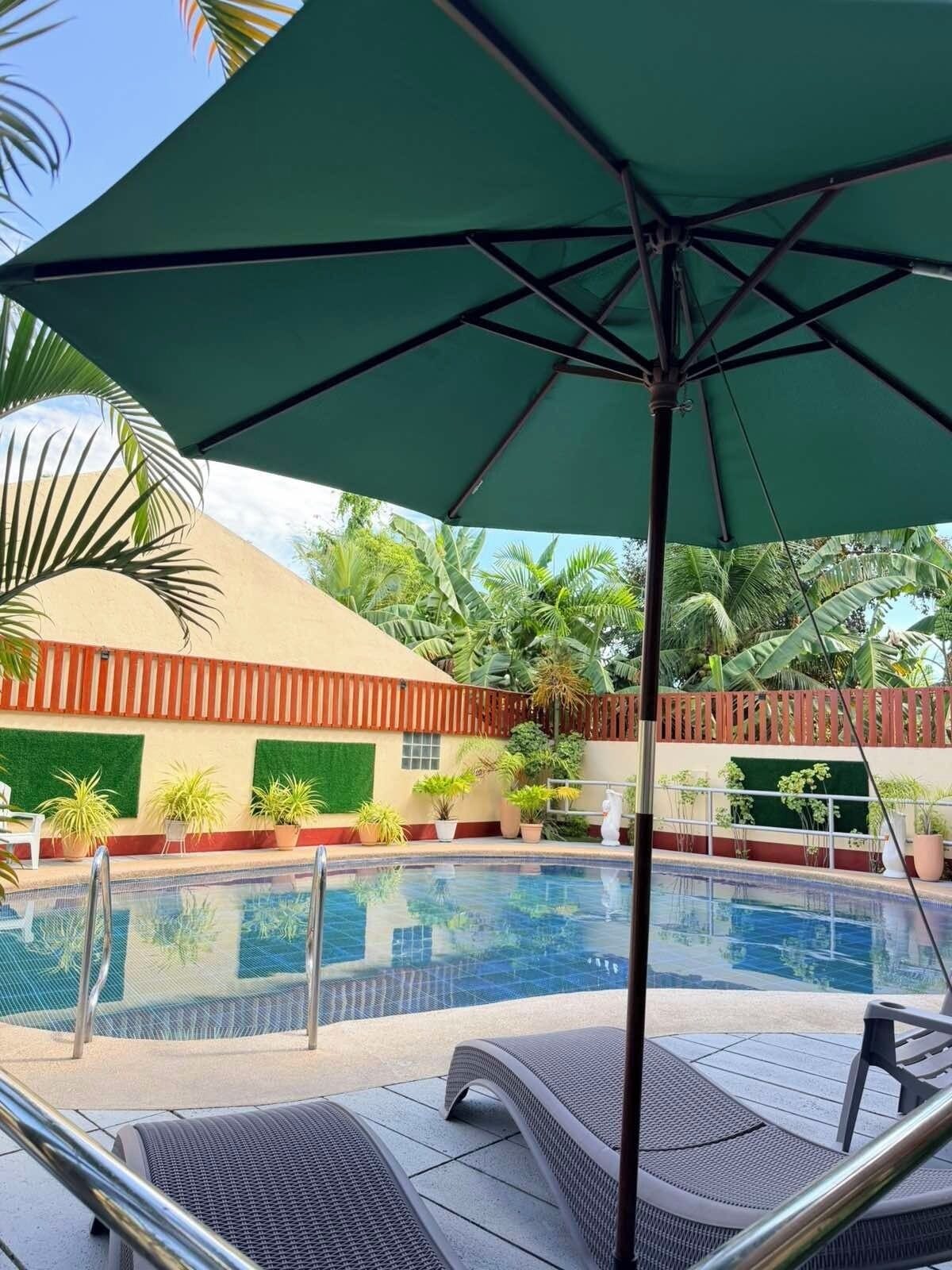 A swimming pool with lounge chairs, a green umbrella, and tropical plants.