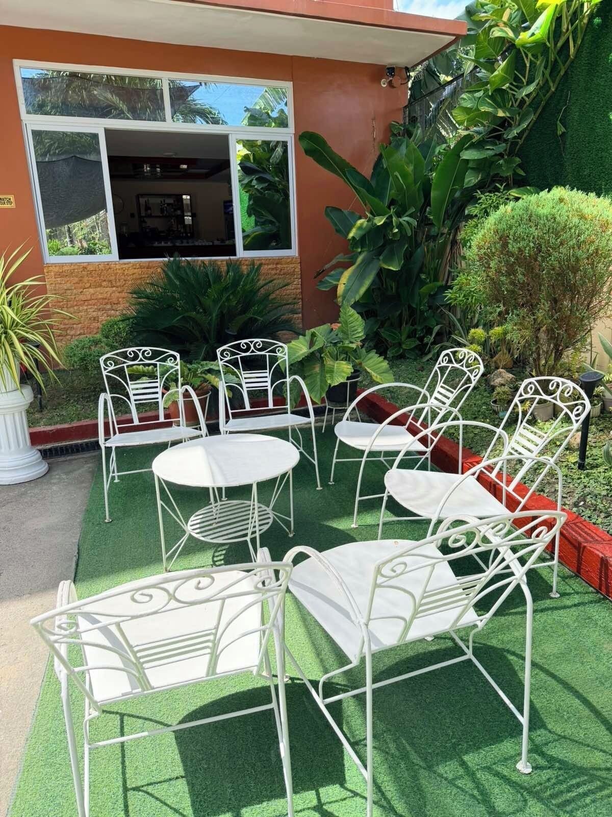 An outdoor seating area with white metal chairs and tables, surrounded by greenery and a building with a window.