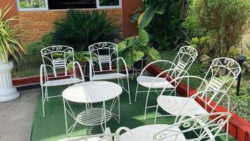 An outdoor seating area with white metal chairs and tables, surrounded by greenery and a building with a window.