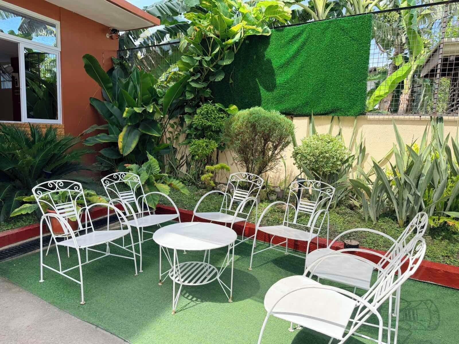 Un patio con sillas y mesas de metal blanco, rodeado de abundante follaje y césped artificial verde.