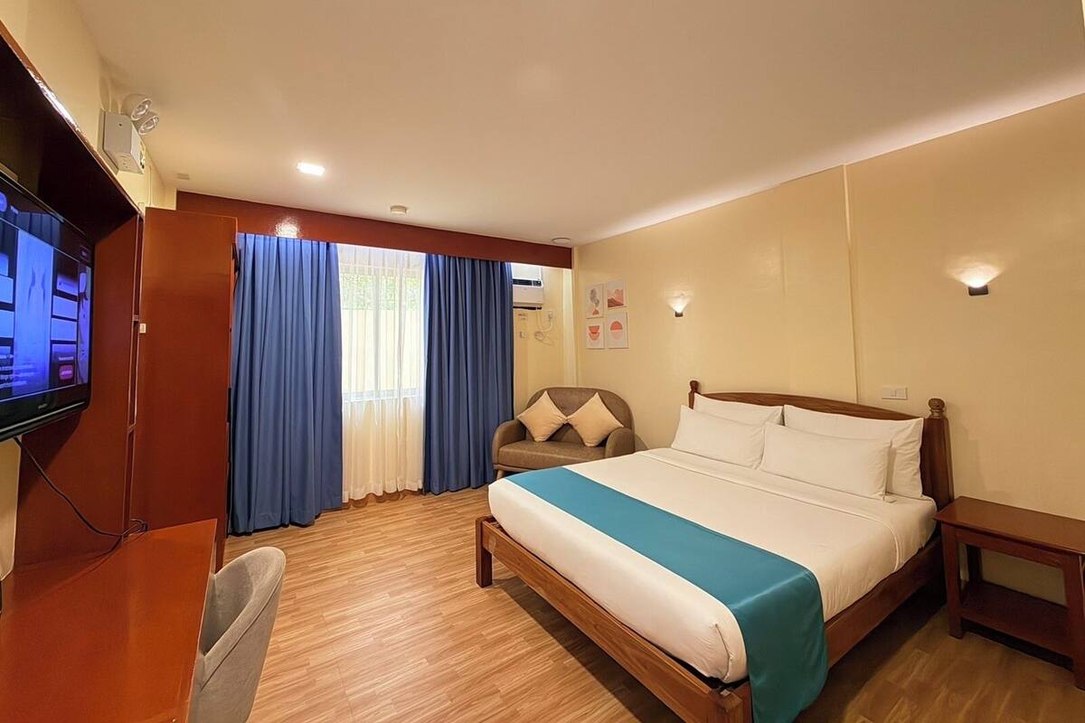 Ein Hotelzimmer mit Bett, Schreibtisch, Stuhl, Fernseher und einem Fenster mit Vorhängen.