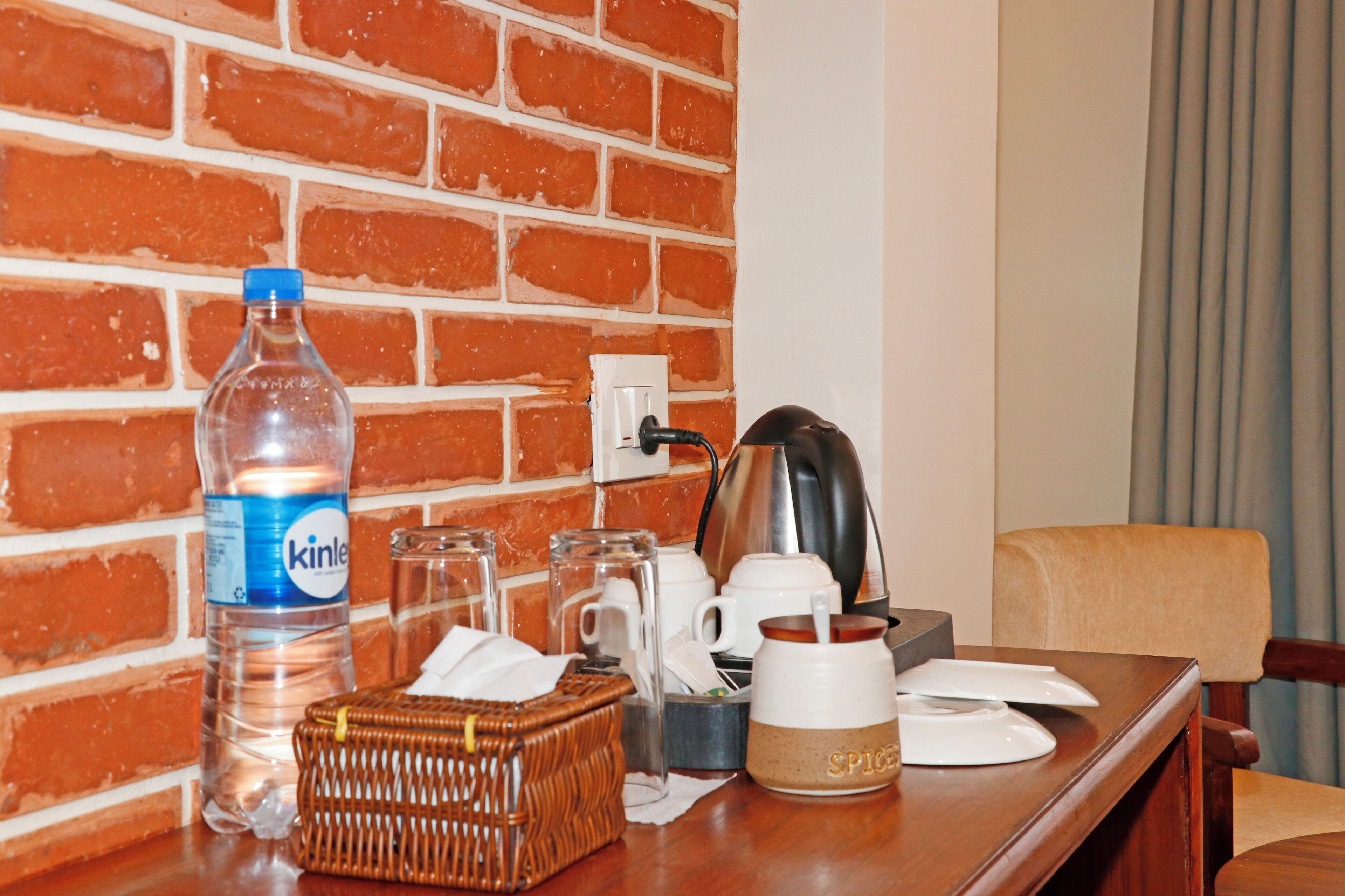 A hotel room with a brick wall, a wooden table with a bottle of water, a kettle, a mug, a basket, and a spice jar.