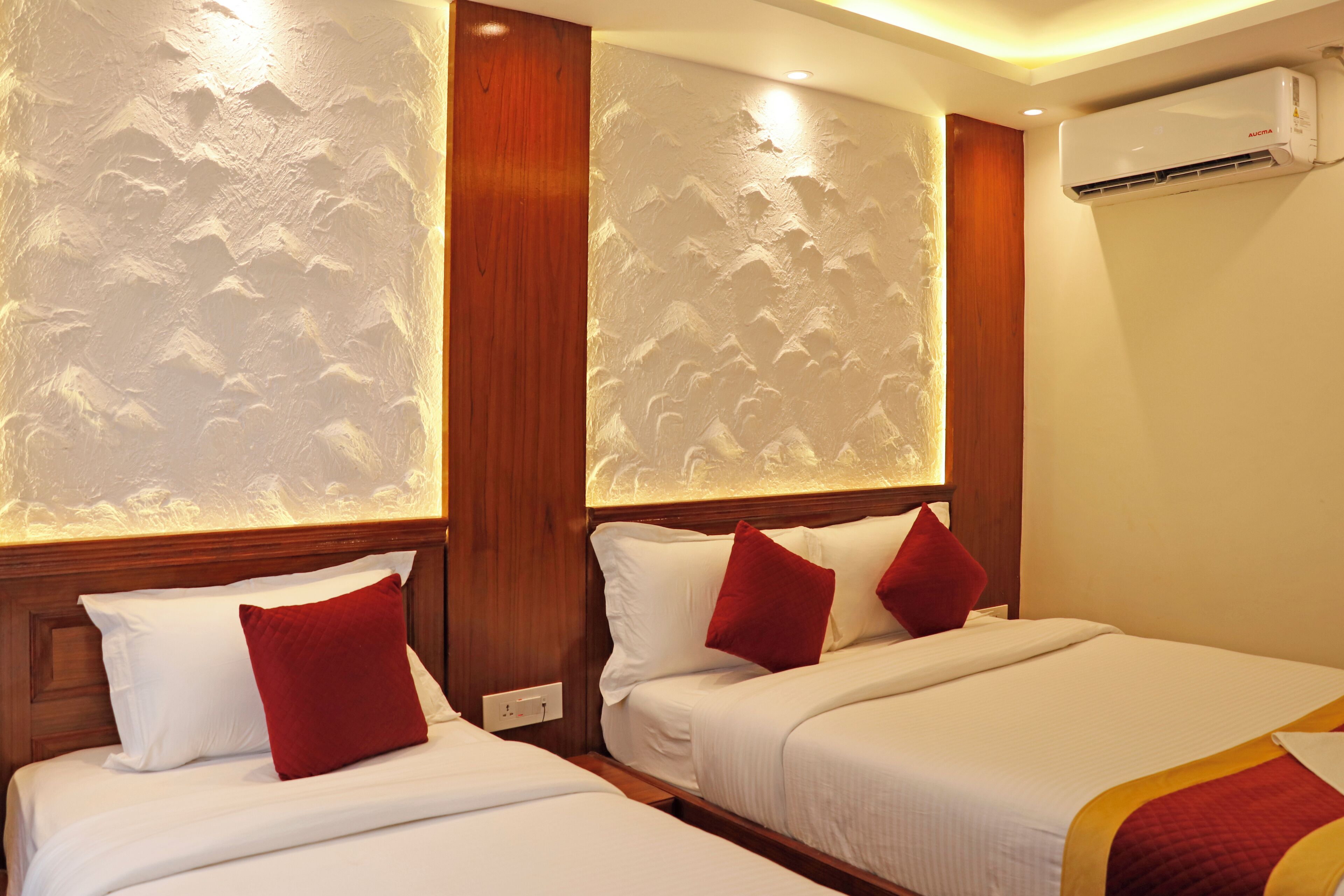 Two beds with white linens and red pillows, a wooden headboard, and a wall-mounted air conditioner.