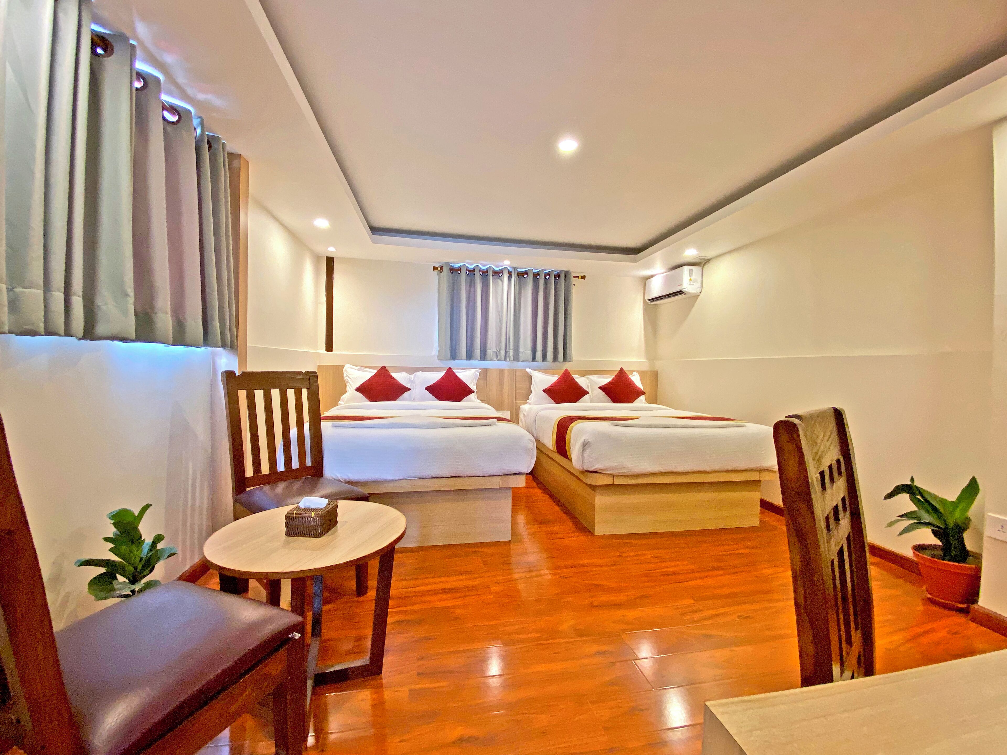 A hotel room with two beds, a small table, a chair, a potted plant, and a window with curtains.