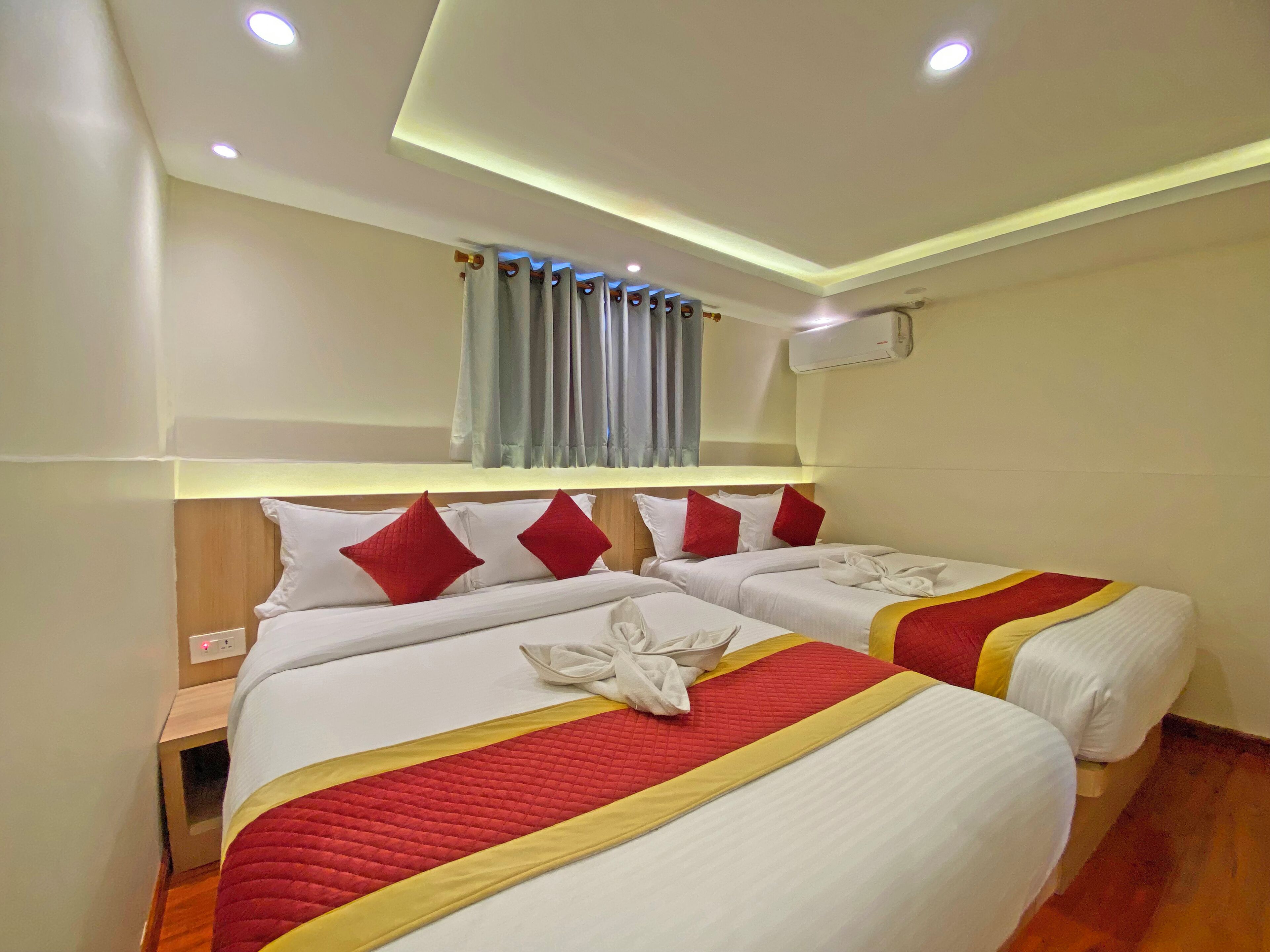 A hotel room with two beds, each with white bedding and red accents, a wooden headboard, a small bedside table, and an air conditioning unit on the wall.