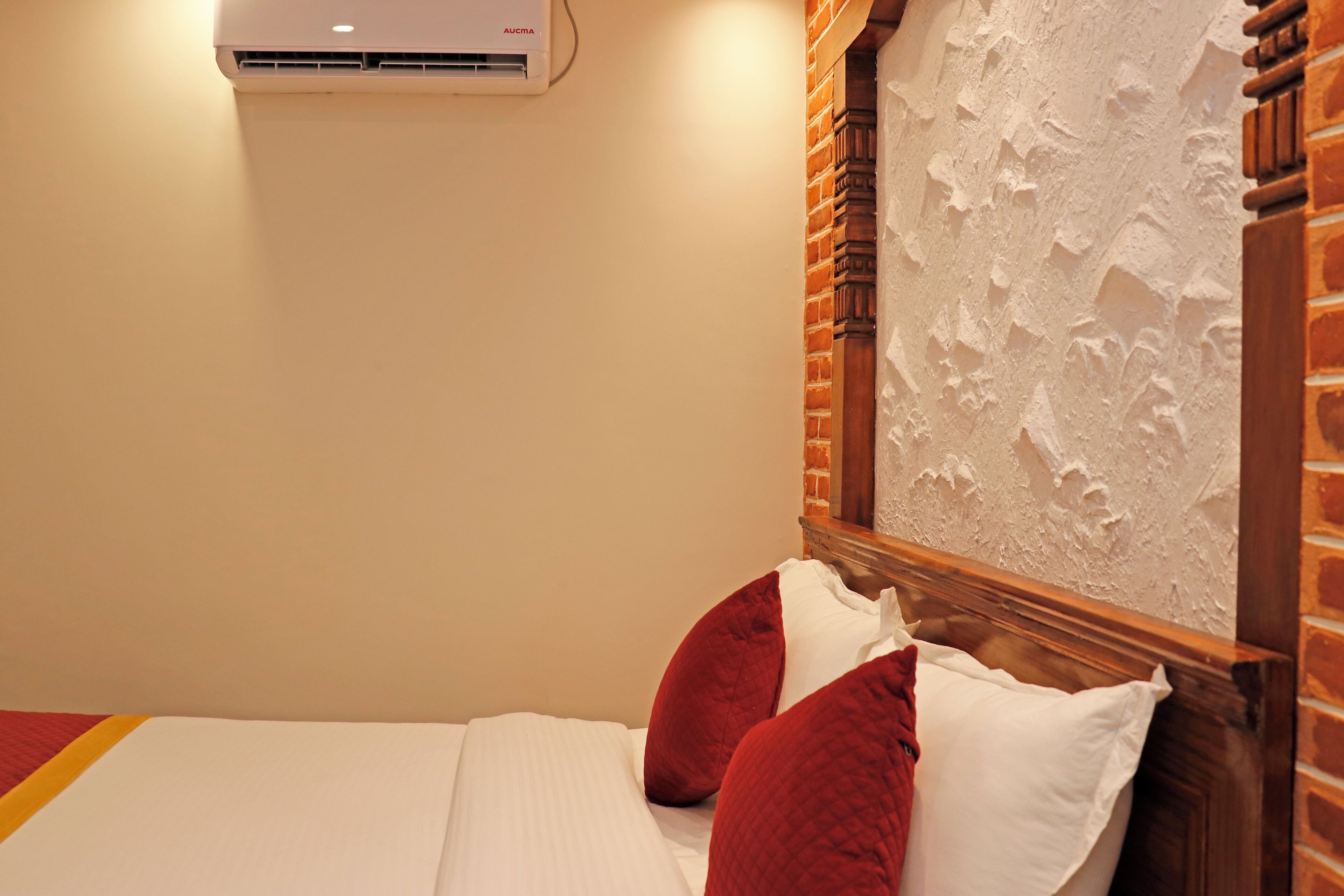 Habitación de hotel con una cama, almohadas rojas y blancas, una pared de ladrillo y un equipo de aire acondicionado.