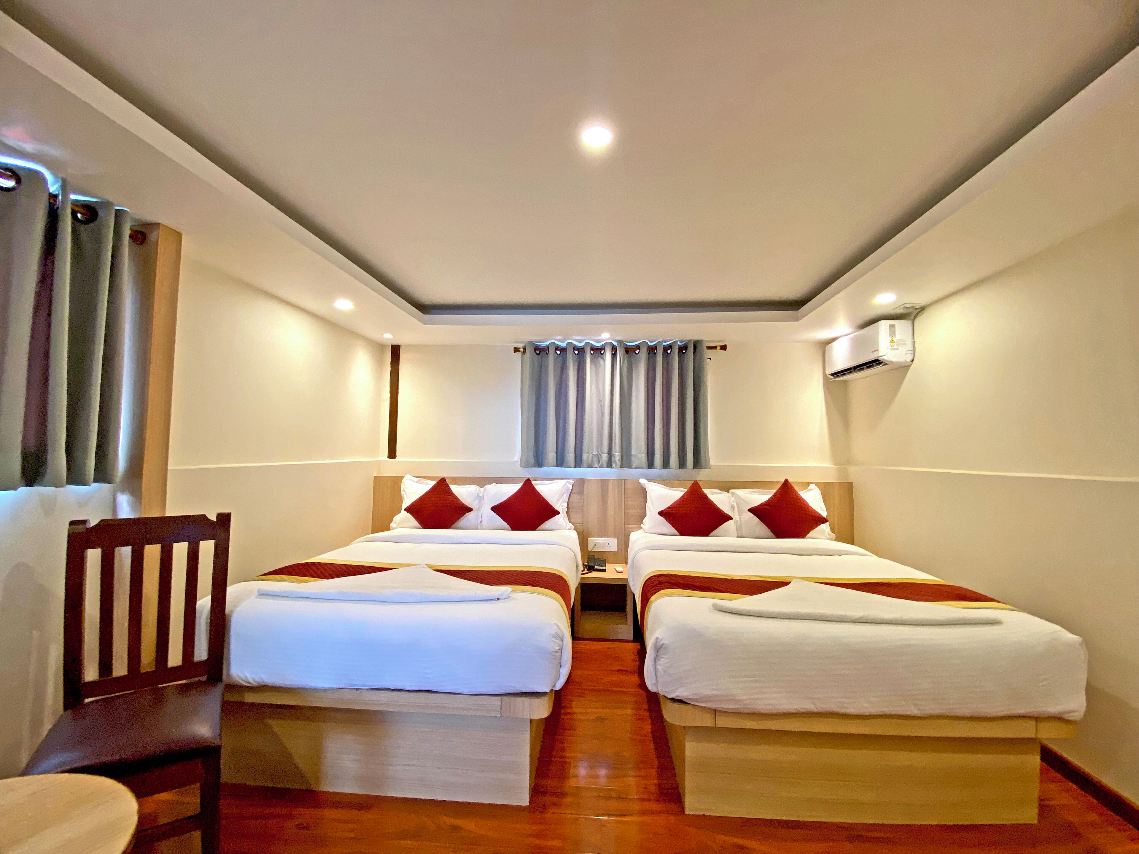A hotel room with two beds, a wooden chair, a small table, and an air conditioning unit on the wall.