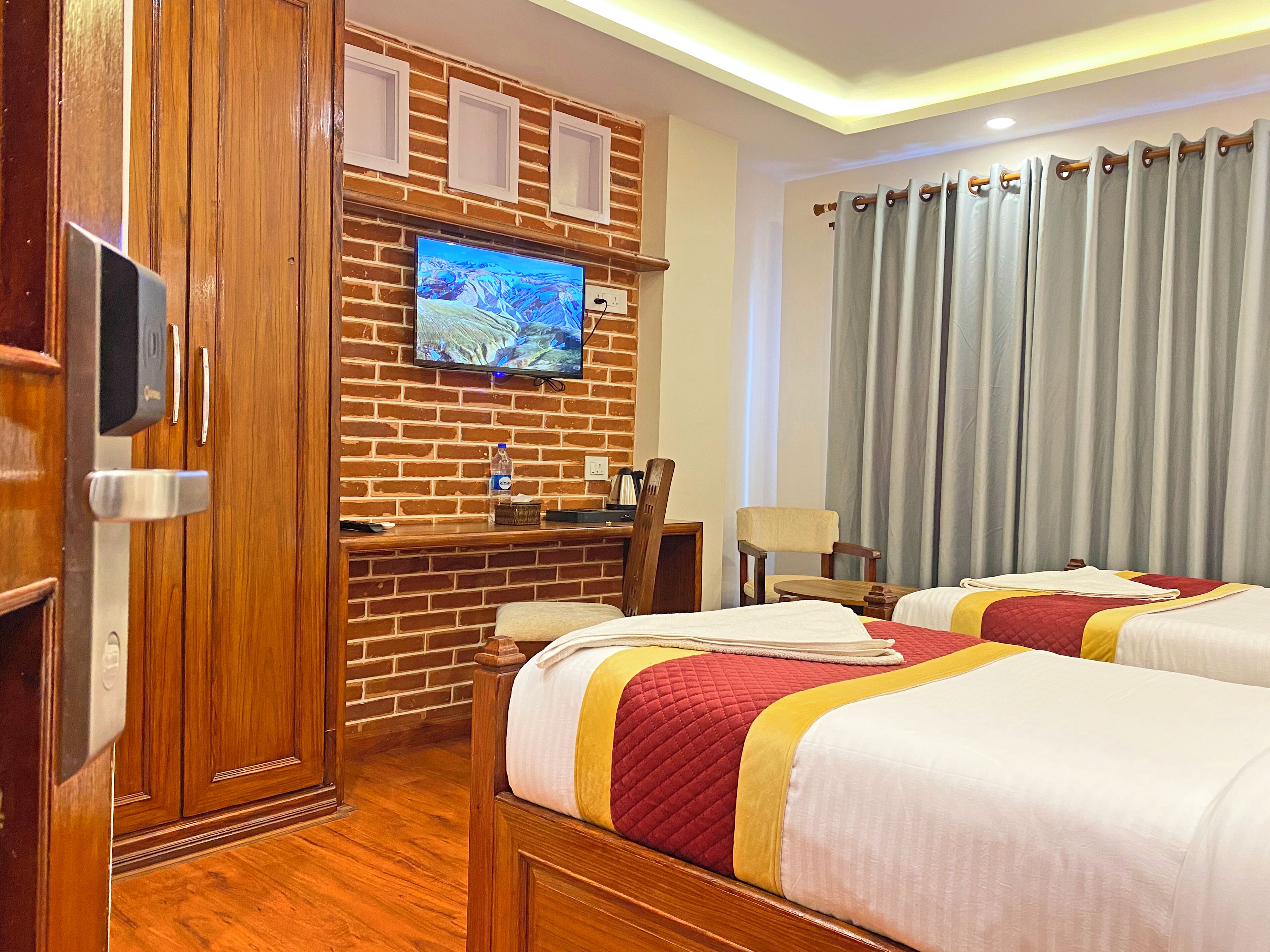 A hotel room with two beds, a TV mounted on a brick wall, a desk with a chair, a window with curtains, and a wooden floor.
