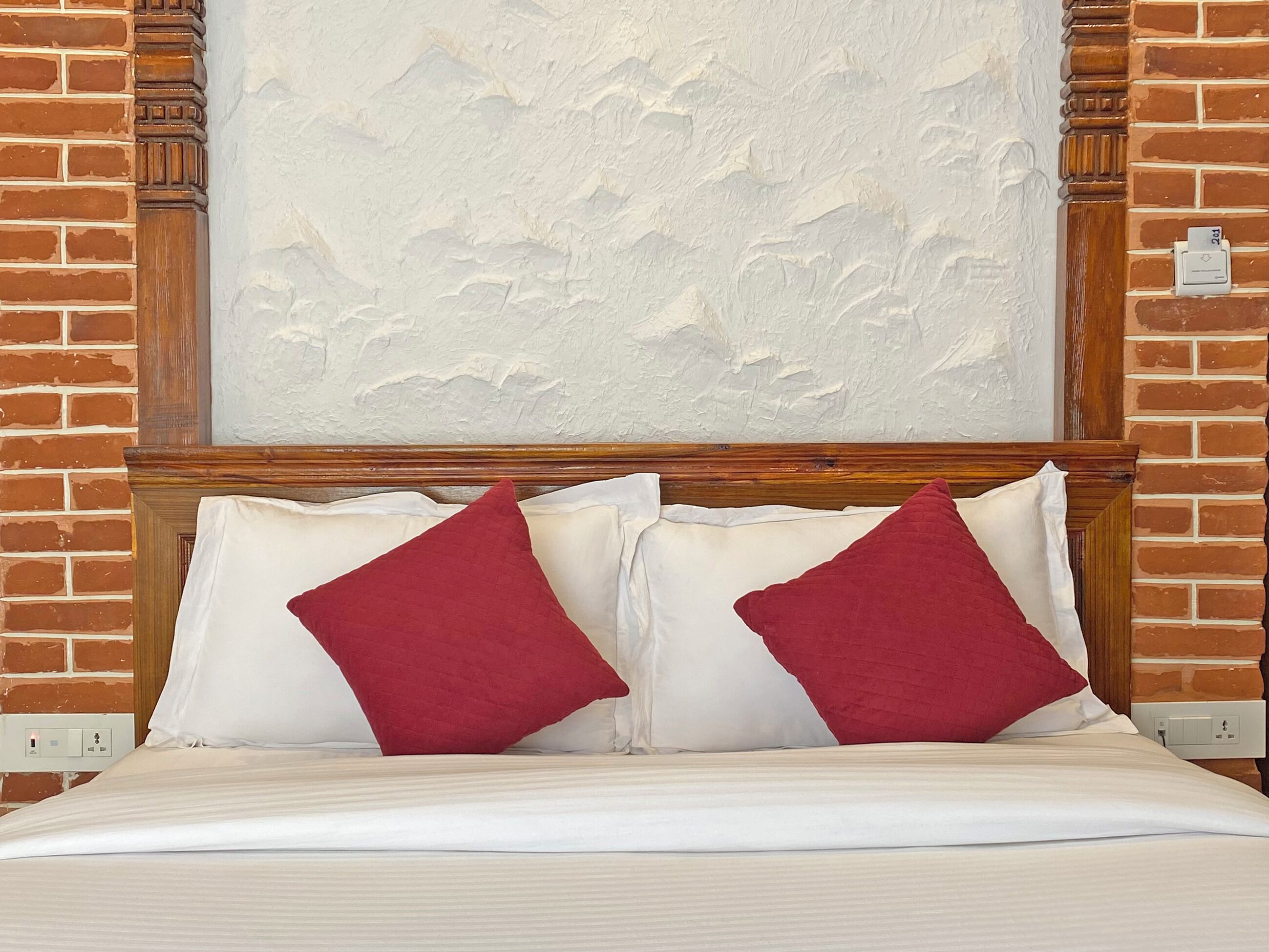 A bed with white bedding and two red pillows, set against a brick wall with a textured white wall above.