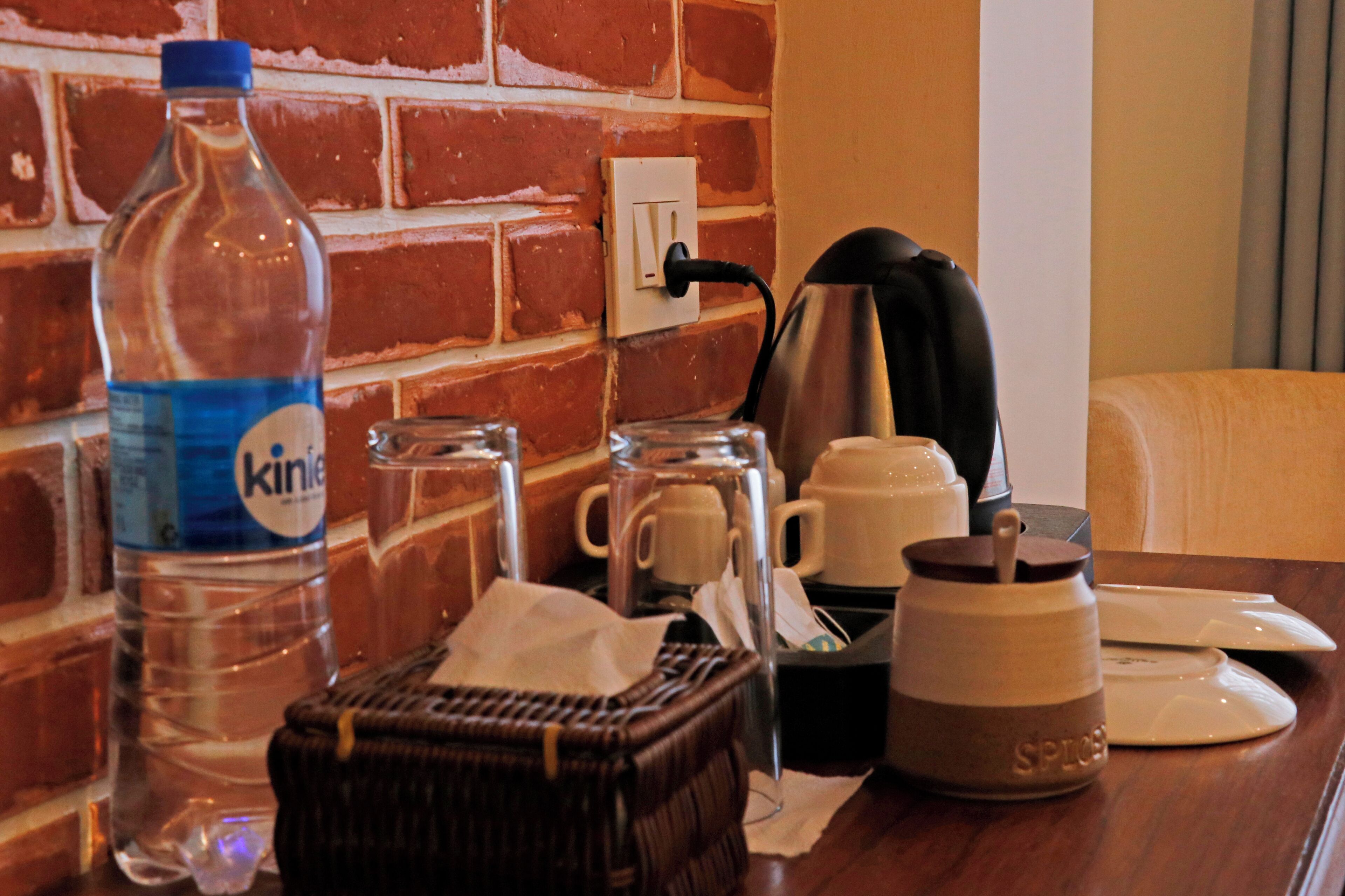 A hotel room with a brick wall, a water bottle, a coffee maker, a mug, a basket, and a small ceramic jar.