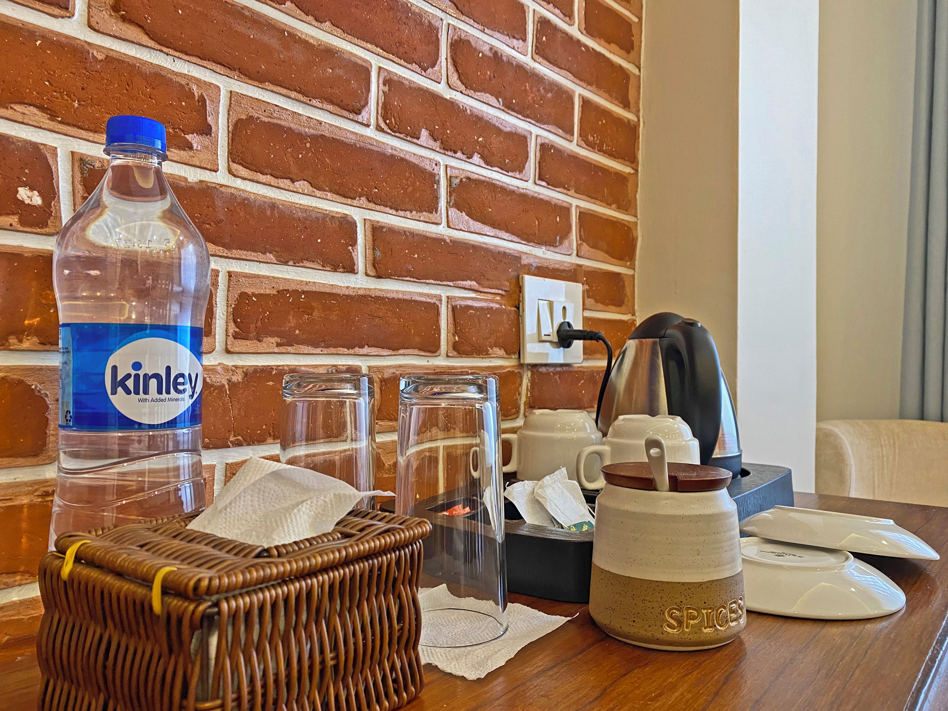 Habitació d'hotel amb paret de maó, ampolla d'aigua, cistella, cafetera i estoig de mocadors.