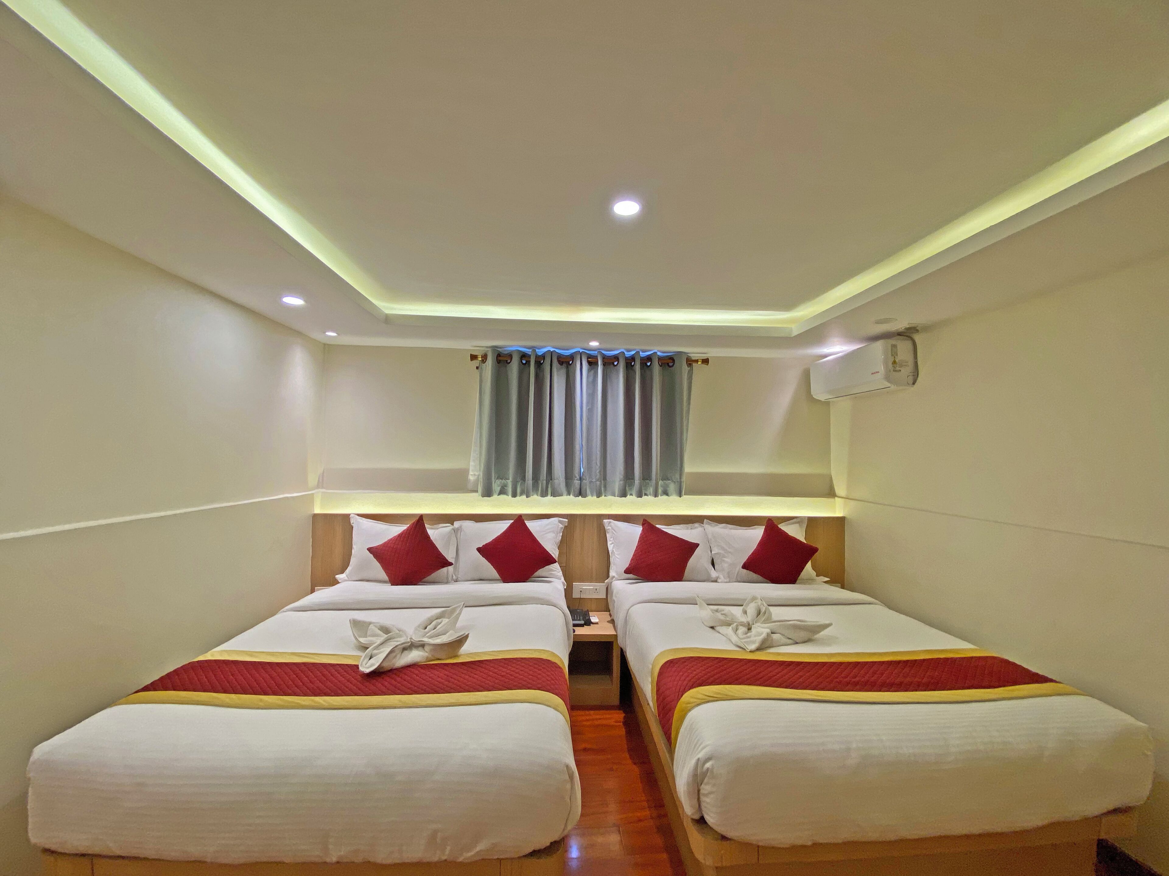 Habitación de hotel con dos camas, cada una con colcha roja y amarilla, almohadas blancas y una toalla blanca sobre ellas. Se ve una ventana con cortinas azules, una lámpara de techo y un equipo de aire acondicionado en la pared.