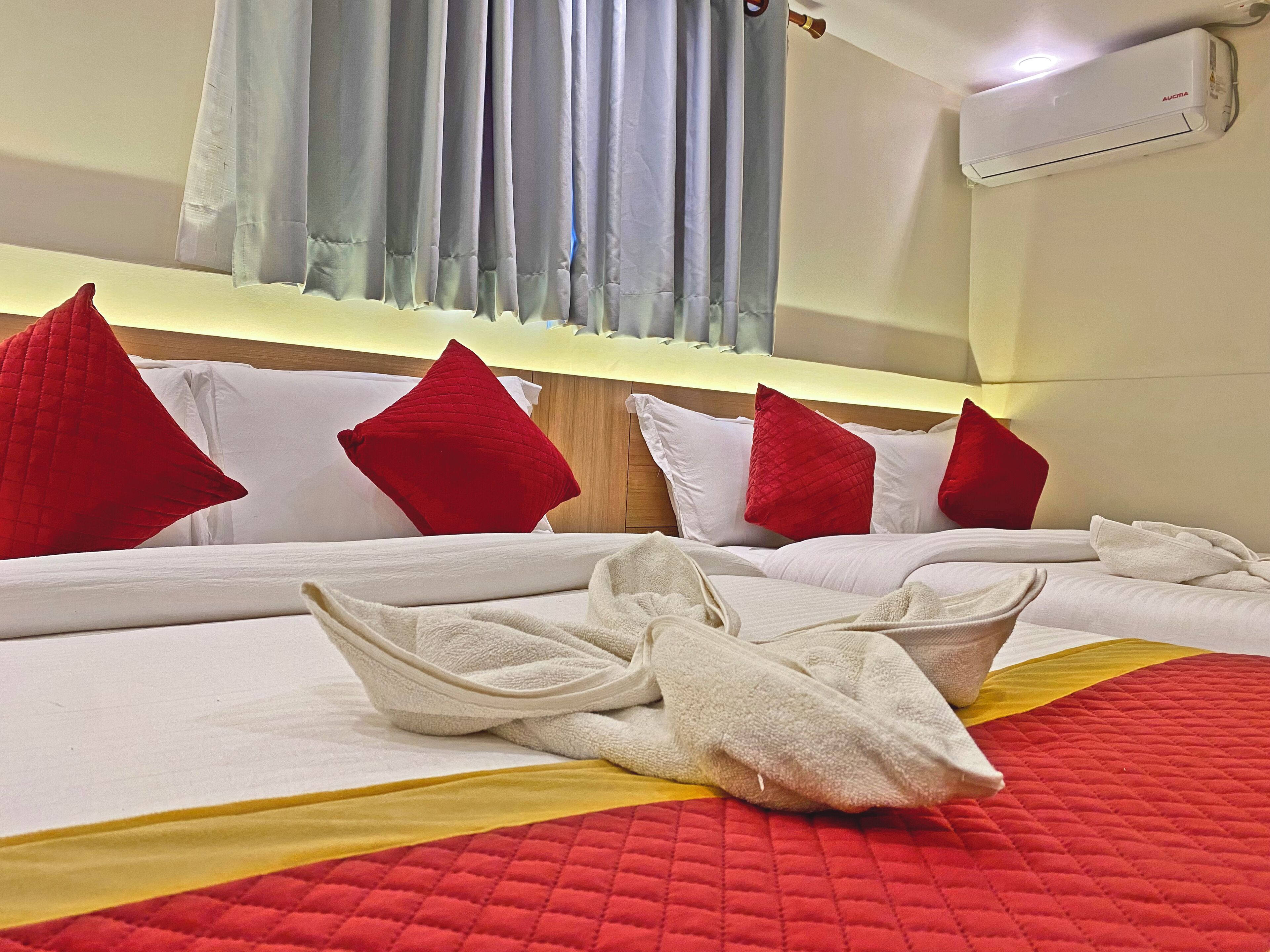 A hotel room with a bed, red pillows, white bedding, a red and yellow quilt, and an air conditioning unit on the wall.