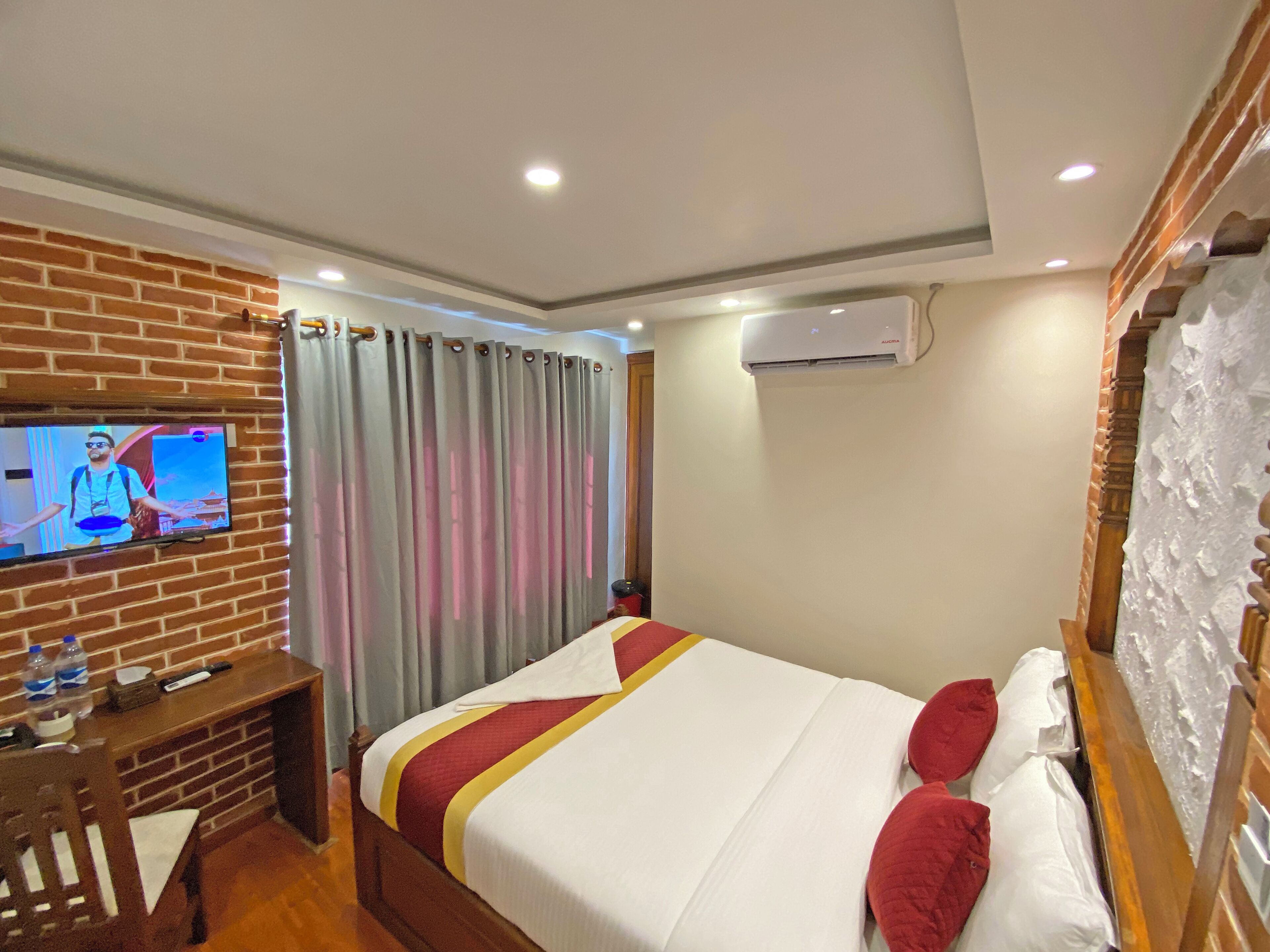 Ein Hotelzimmer mit Fernseher an einer Ziegelwand, einem Bett mit rot-weißer Bettwäsche, einem Nachttisch aus Holz und einer Klimaanlage an der Wand.
