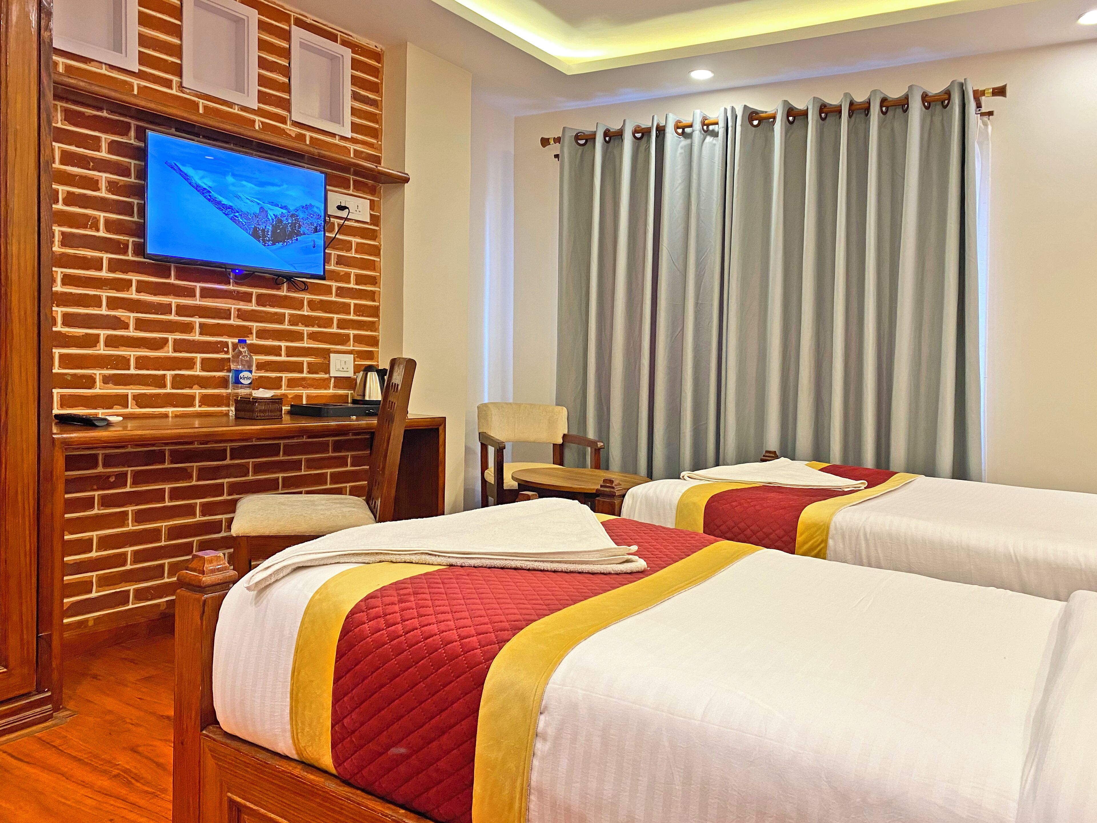 A hotel room with two beds, a TV mounted on a brick wall, a desk with a chair, and a large window with curtains.