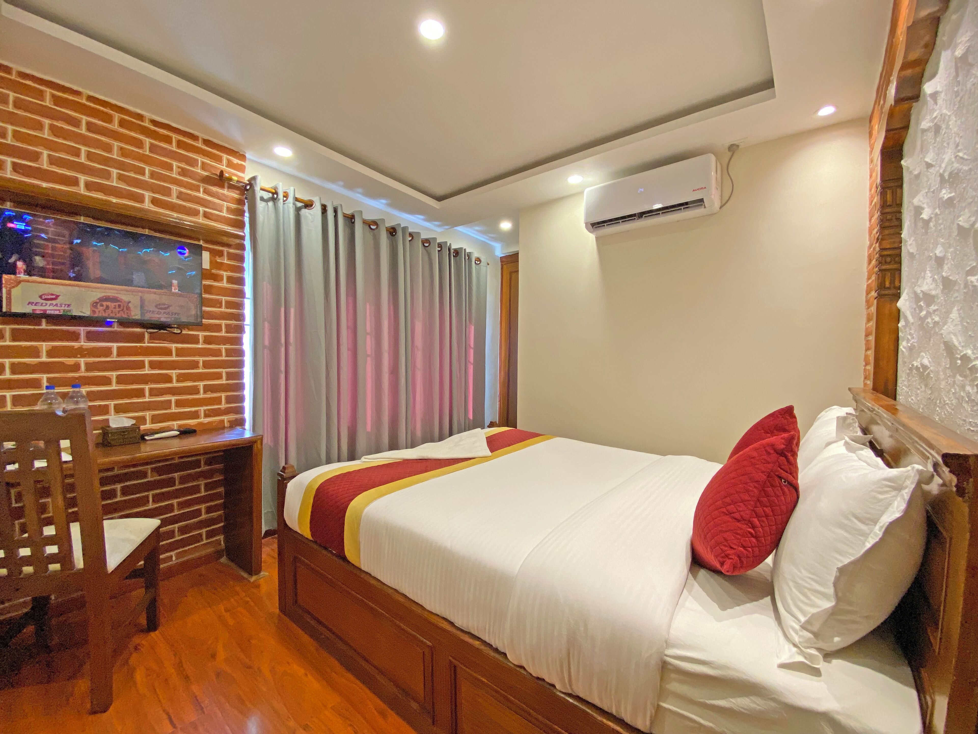 Une chambre d’hôtel avec un mur en briques, un téléviseur fixé au mur, un lit avec une literie blanche et des oreillers rouges, un bureau en bois avec une chaise et un appareil de climatisation fixé au mur.