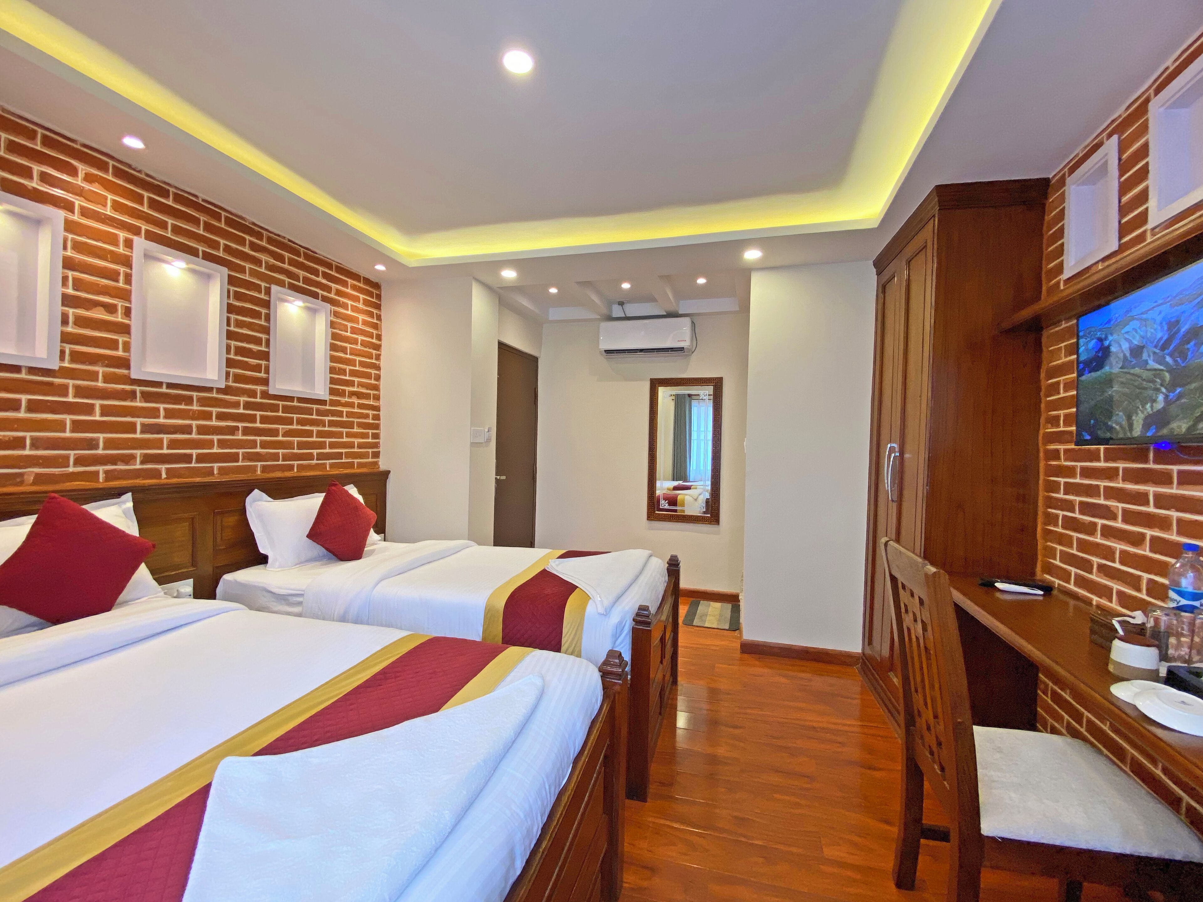 A hotel room with two beds, a TV, a wooden desk, and a brick wall.