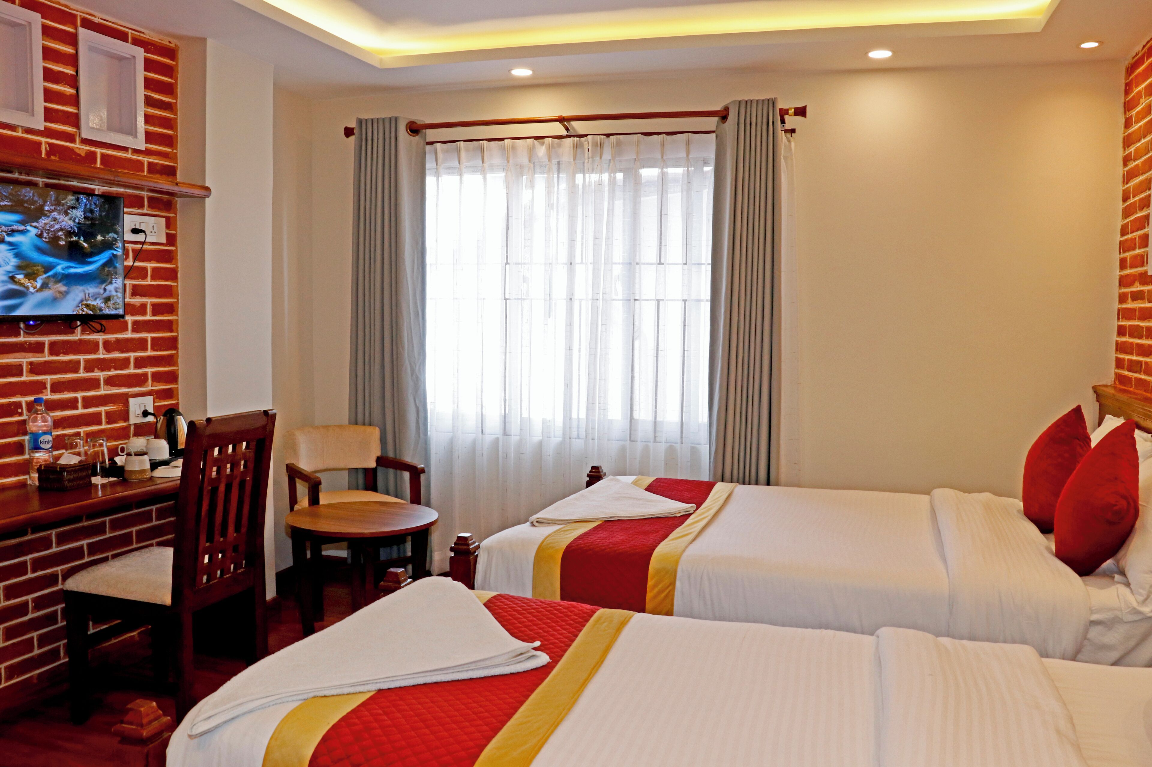 A hotel room with two beds, a TV mounted on a brick wall, a desk with a chair, and a window with curtains.