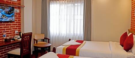 A hotel room with two beds, a TV mounted on a brick wall, a desk with a chair, and a window with curtains.