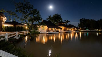 A waterfront property at night with reflective water, a wooden dock, and several illuminated buildings.