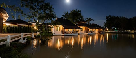 A waterfront property at night with reflective water, a wooden dock, and several illuminated buildings.