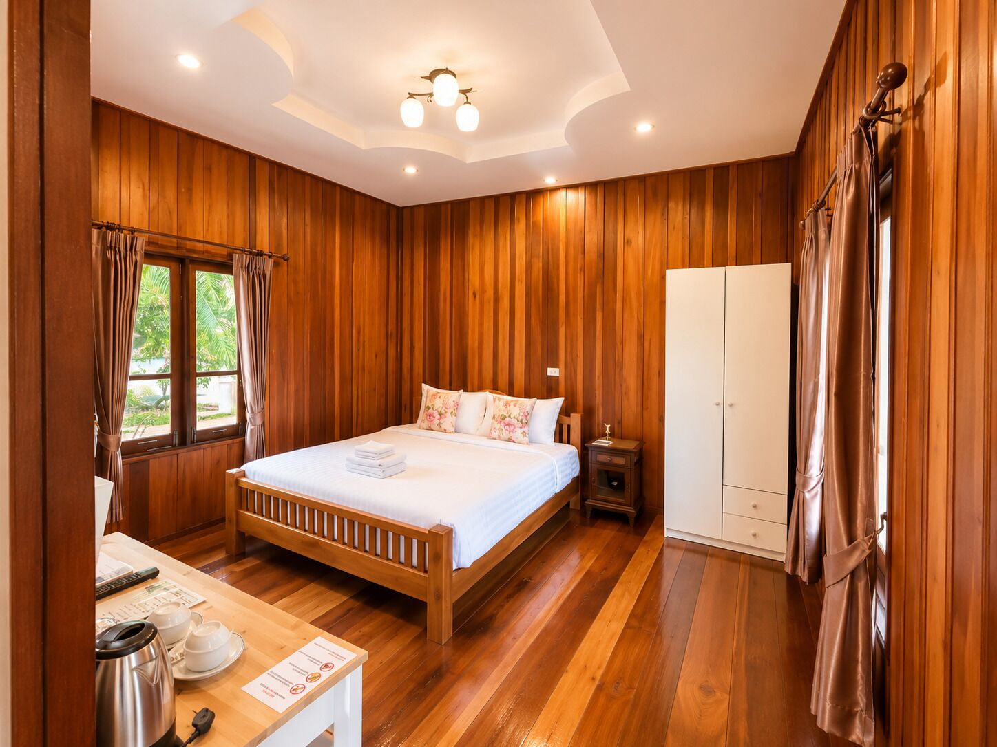 A wooden-paneled room with a bed, a wardrobe, a table with a kettle, and a window with curtains.