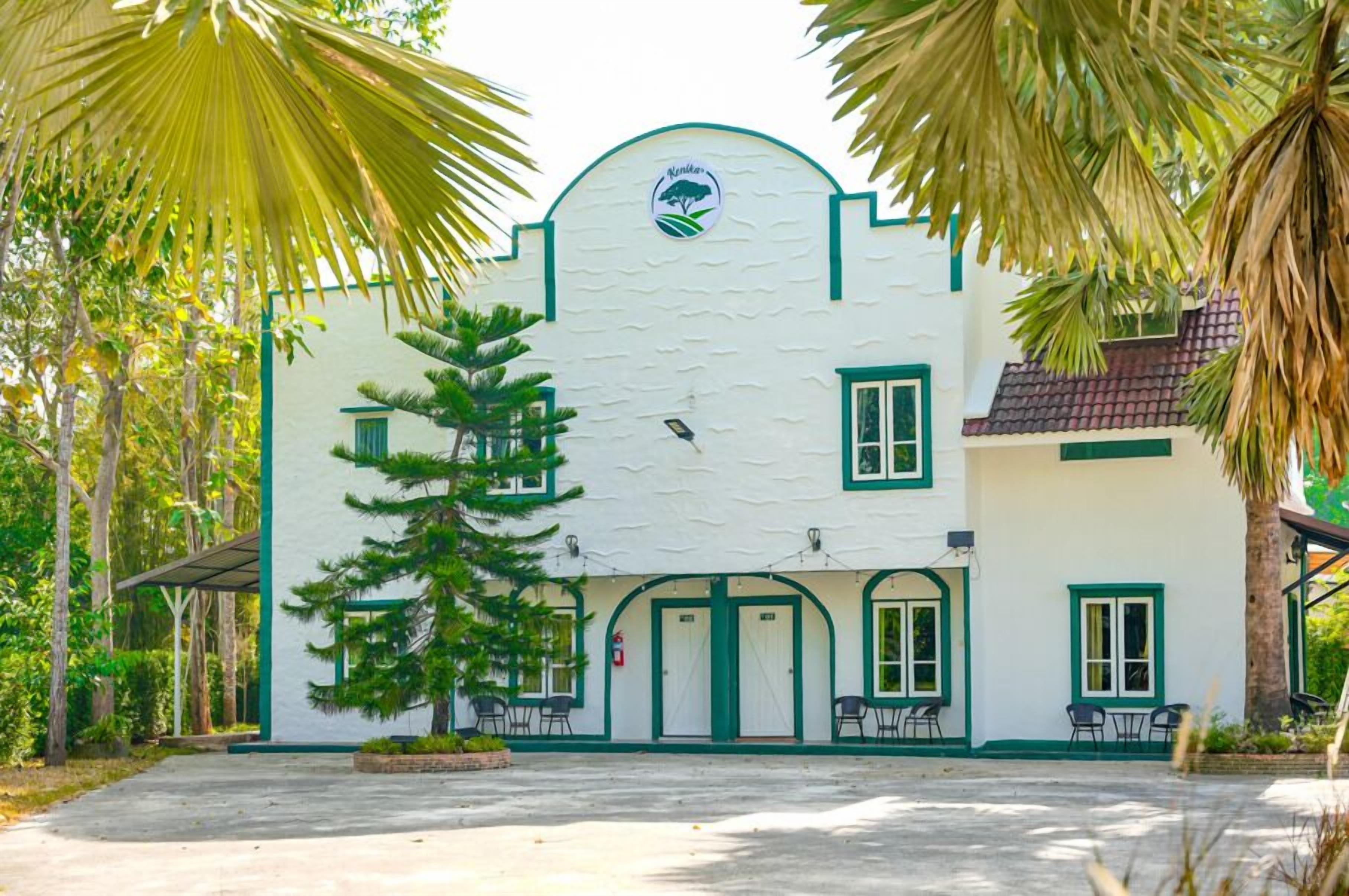Um edifício com fachada branca, detalhes em verde e um logotipo com uma árvore. Há várias portas e janelas, e algumas cadeiras são visíveis do lado de fora.