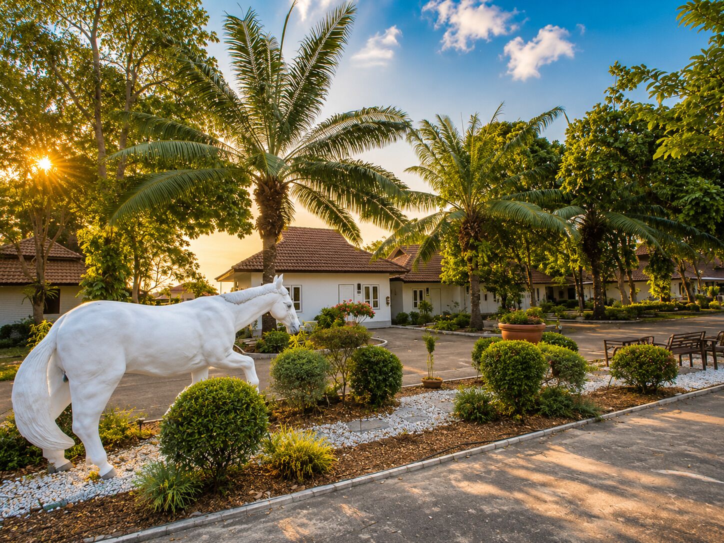 Una estatua de un caballo blanco en un jardín ajardinado con palmeras y un edificio al fondo.