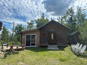 Une cabane en bois avec une porte d’entrée, une porte-fenêtre coulissante et un petit porche équipé de chaises.