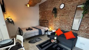 A compact living space with a bed, a sofa with orange cushions, a dining table with plates, and a small side table with a bottle and a bowl.