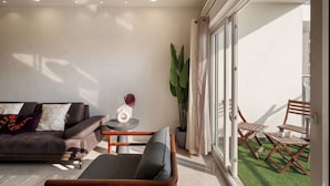 A modern living room with a sofa, a small table, and a chair. There is a large window with curtains and a view of the outside.