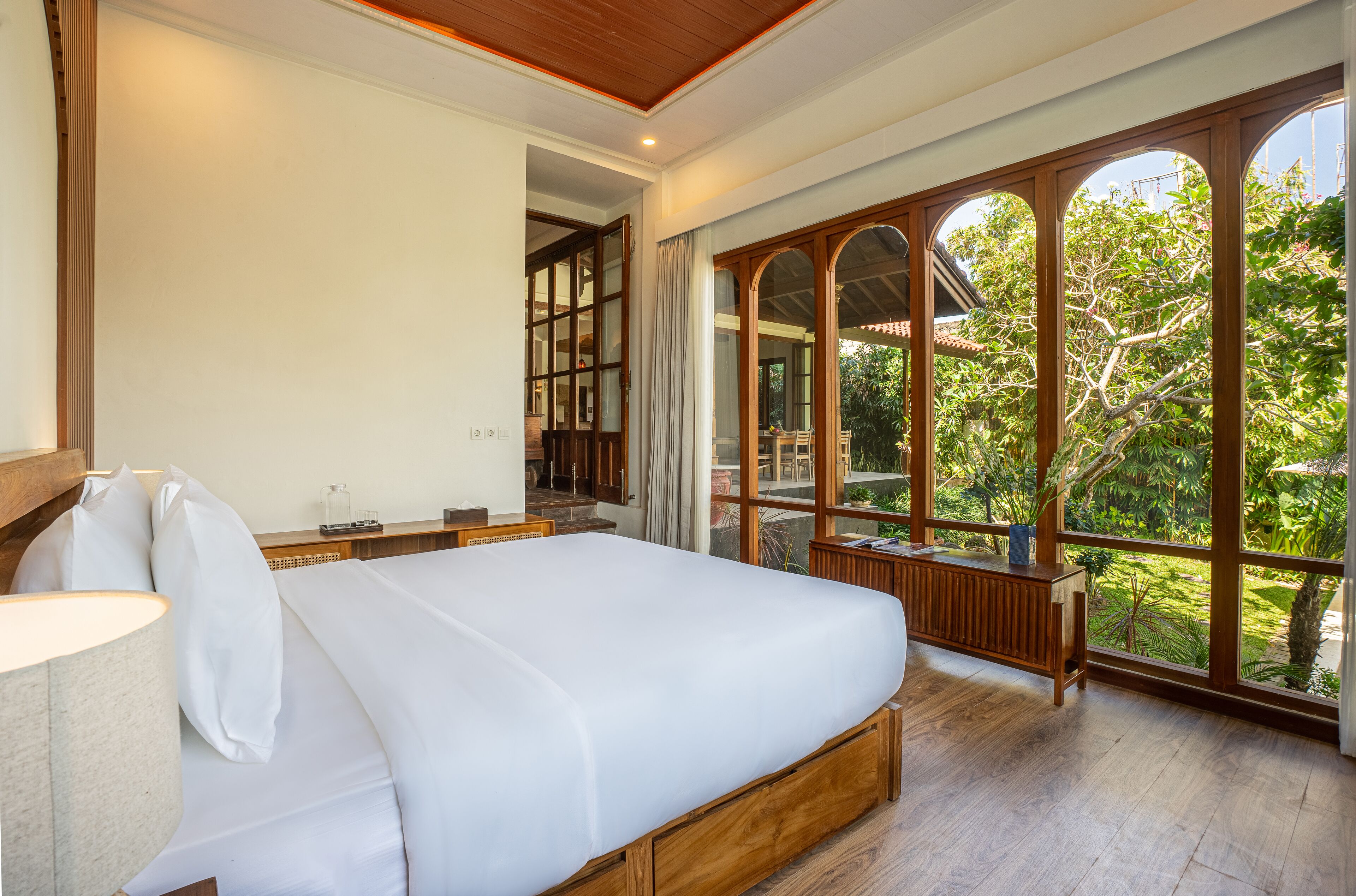 A bedroom with a large bed, wooden headboard, and a view of a lush garden through arched windows.
